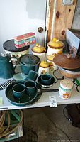 Wide shot showing ceramic mushroom canisters, green speckled cookware including two cups, a frying pan, covered pot, coffee percolator, and decorative tins on table