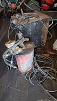 Photo showing the vintage Craftsman arc welder with black metal housing and attached cables alongside a rusted red metal container with attached hoses and valves covered in grime and dust.