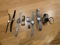 All 9 watches laid out on wooden floor, variety of men's and women's watches with different band styles and faces.