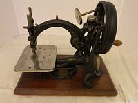 Front side view of small cast iron sewing machine on wooden base, showing hand crank and sewing plate