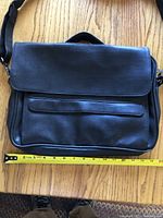 Closed black leather briefcase showing front with external zipper pocket and tape measure at base indicating width around 16 inches.