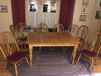 Set of oak dining table and 8 matching oak chairs arranged in dining room, showing table's rectangular top, turned legs, and chairs with spindle backs and burgundy seat cushions.