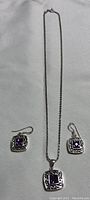 Silver pendant necklace with matching pierced earrings laid flat on grey fabric background. The necklace chain is thin silver. Pendant and earrings have square purple amethyst stones in ornate silver settings.