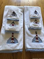 Two sets of folded white towels with lighthouse motifs arranged side by side on a wooden surface. Towels include bath towels, hand towels, and washcloths with consistent embroidered lighthouse design.