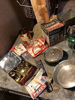 Photo showing brass camping stove in red metal case with fuel packets and small aluminum pot with lid on table.