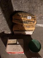 Two bags of Quikrete Concrete Mix stacked against a concrete wall with a box and green bucket on the floor.