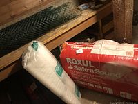 Photo of Roxul Safe'n'Sound insulation package and roll of white construction material with green wire mesh behind.