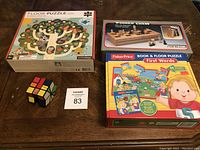 Photo of lot showing two floor puzzles, one wooden chess game in box, and a Rubik's Cube on table.