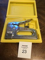 Metal heavy duty stapler with 'Heavy Duty' label inside a yellow plastic carrying case on wooden surface. Auction lot number card included in image.