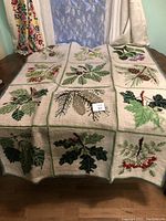 Full view of rectangular needlepoint textile on table, showing nine embroidered botanical square panels including leaves, berries, and birds.