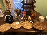 Various household items arranged on a wooden table including woven baskets, glassware, metal and ceramic containers, copper pot, and Coca-Cola collectible