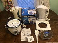 Black & Decker rice cooker with box, two white electric kettles, lid, and spoon on table.
