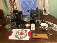 Wide view of assortment of kitchen items on table showing pots, canisters, cutting boards, enamel bowls, grinder, and rolling pin.