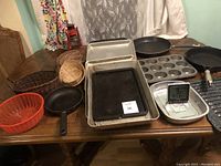 Wide view of all kitchen items on wooden table including bakeware, cookware, plastic bowl, and woven baskets