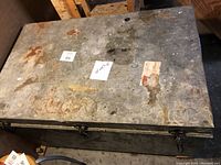 Top view of large rectangular metal box showing worn lid surface, rust spots, and stickers.