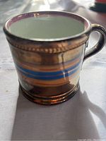 Close-up image of the cup showing the lustreware gold finish and blue and light brown bands near the center. White unglazed interior is visible.