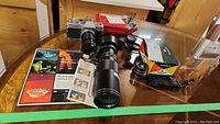 Camera with large lens attached, flash, additional lens, original flash box, and pamphlets on a glass tabletop.