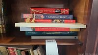 Books stacked on a shelf including Elusive Destiny, Who in Canadian Sport, Les Désirent Une Patrie Meilleure, The Canadian Establishment, Kar/Ha and others.