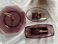Three Moroccan amethyst glass serving trays with gold-tone handles arranged on white cloth.
