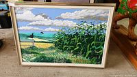 Full view of the framed painting depicting a rural landscape with tall green corn stalks, birds on a post, fields, hills, and a cloudy blue sky.