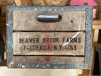 Front side of the wooden milk crate showing "BEAVER BROOK FARMS 7- ITHACA N.Y. 53" stencil in black text, with metal corner reinforcement and wood handles.