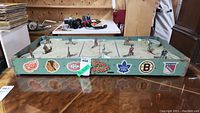 Full view of the vintage table hockey game showing frame and player figures positioned on the rink surface, with visible logos of Original Six NHL teams on the sides.