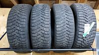 Four used winter tires placed side by side on a wooden shelf rack, showing tread and general condition.