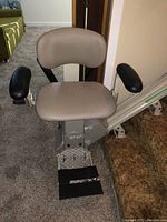 Photo of the electric stairlift chair showing beige padded seat, brown armrests, and metallic base with footrest.