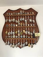 Front view of a wooden display plaque holding 36 collectible souvenir spoons in four rows with various colored and metallic finishes.
