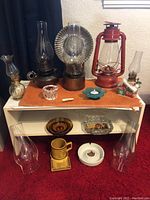 Photo of assorted vintage oil lamps and lanterns on top of a small white shelf unit, including small clear glass oil lamp, tall dark glass oil lamp, large clear ribbed glass oil lamp, and red metal hurricane lantern. Various ashtrays and a ceramic souvenir house also visible.