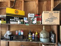 Photo showing several oil cans, automotive fluids, funnels, and a large translucent plastic container on wooden shelves, with Gulf Auto Diesel metal sign and a box labeled 'Transmission Power Steering Fluid'