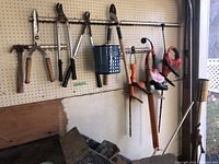 Wide view of assorted garden trimmers and cutting tools hanging on pegboard, showing 3 orange/red electric trimmers and multiple manual trimmers including hedge shears, loppers, and pruning shears.