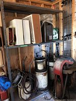 One metal shelving unit holding four portable electric heaters and fans on the top shelf and two white cylindrical oil drainer containers on the bottom shelf.