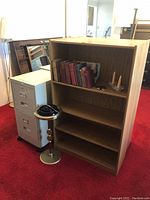 Wooden bookshelf, metal filing cabinet with two drawers, and brass-colored stand-up ashtray with wooden accents placed on red carpet.