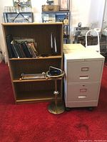 Wooden bookshelf with books, cream-colored filing cabinet, and stand-up ashtray grouped on red carpeted floor.