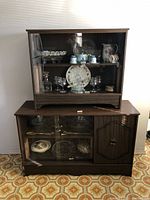 Two stacked wooden cabinets with glass sliding doors; one smaller on top and one larger below with a wooden door panel. Cabinets contain no items inside.