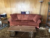 Wide view showing the red fabric couch with cushions, coffee table in front, and floor lamp to the right against wooden wall paneling.