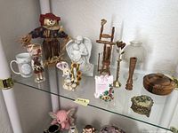 Glass shelf displaying a vintage collectibles assortment including porcelain angel, scarecrow, Scottish doll in tartan, brass bell, miniature furniture, and various decorative items.