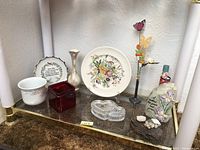 Overview shot of lot showing assortment of vases, decorative plates, glass containers, and figurines on a shelf with textured surface.