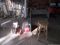 View of the vintage tiller with pneumatic tires, compressor, metal table, and wooden stool in garage setting