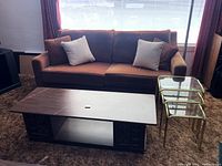 Wide shot showing brown vintage fabric couch with square pillows, rectangular wood coffee table in front, and glass nesting tables with gold frames to the side