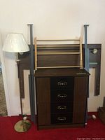 Photo shows dresser with four drawers featuring brass handles, floor lamp with brass base and white shade, and disassembled wood bed frame parts leaning against the wall.