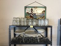 Wide shot of metal shelving holding clear glasses on top shelf, plates and bowls on lower shelves with stained glass picture hanging on wall above.