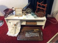 White wooden shelf with various items including wooden well, brass ship wheel, owl figurine, glass fishing floats, cutlery set in wooden box, angel figurine, cutting board with knives and utensils, and small wooden wheelbarrow.