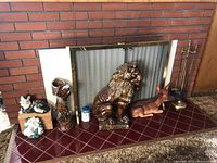 Porcelain animal figurines and fireplace tools displayed on a red tiled hearth in front of a fireplace with a brass mesh screen. Items include a black and white cat figurine, brown glazed dog head statue, large brown lion, brown deer, brass fireplace tools on stand, white and blue lighthouse decorative piece.