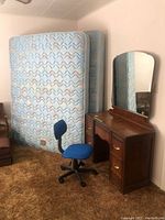 View of queen size mattress and box spring leaning against the wall with vintage wooden desk and blue office swivel chair in front.