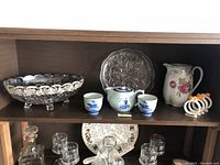 Wide view of the shelf showing the crystal bowl, ceramic pitcher, silver napkin holders, blue and white ceramic tea set and decorative plate