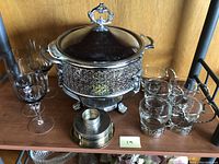 Photo of silver plated serving bowl with lid and stand on shelf with glass cups and silver plated coasters in front