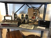 Photo of vintage electronics on metal shelves, including cordless phones, a wooden and metallic electronic item, film reel, and black RCA alarm clock.
