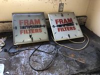 Two square FRAM Filters clocks side by side showing front faces on a workbench